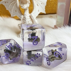 May include: Four clear purple dice with black cat illustrations and purple numbers. The dice are on a white fluffy surface.