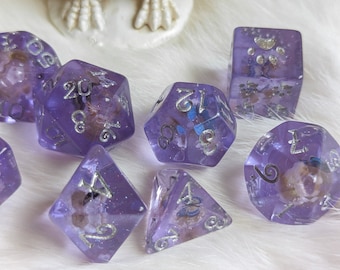 Puede incluir: Un conjunto de siete dados morados con números blancos y purpurina plateada. Los dados son translúcidos y tienen una variedad de formas, incluyendo un d4, d6, d8, d10, d12 y d20.