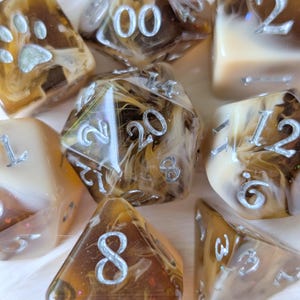 Könnte beinhalten: Eine Sammlung von Würfeln in Braun-, Beige- und Cremetönen mit silbernen Zahlen. Einige Würfel haben einen wirbelnden, marmorierten Effekt. Ein Würfel hat ein Pfotenabdruck-Design. Für Tischspiele.