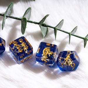 May include: A set of four blue resin dice with gold-colored designs. The dice have a celestial theme, with paw prints, stars, and other symbols. The dice are displayed on a white, fluffy surface with a green leafy branch.