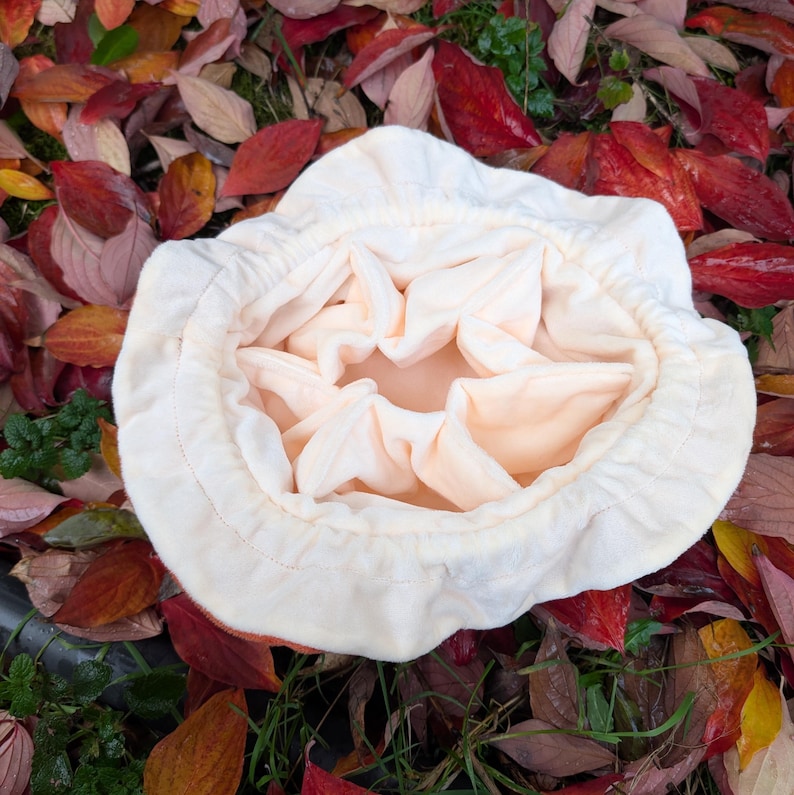May include: A cream-colored, soft fabric hat with a gathered interior, resting on a bed of colorful autumn leaves. The hat's brim is slightly ruffled, and the interior fabric is a light peach color.