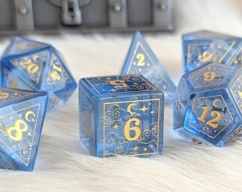 Könnte beinhalten: Set aus sieben blau-goldenen Würfeln mit Himmelsmotiven. Die Würfel haben goldene Zahlen und komplizierte Muster aus Sternen und Monden. Die Würfel bestehen aus einem durchscheinenden Material.
