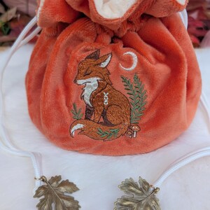 May include: An orange velvet drawstring bag with an embroidered fox design. The fox is brown and white, with a crescent moon and foliage. The bag has a white drawstring and leaf-shaped charms.