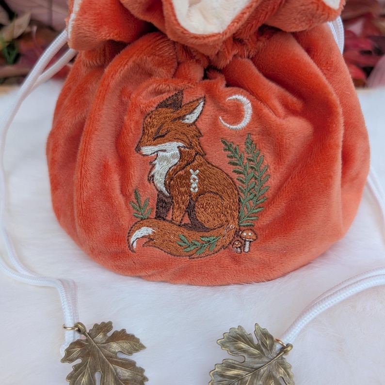 May include: An orange velvet drawstring bag with an embroidered fox design. The fox is brown and white, with a crescent moon and foliage. The bag has a white drawstring and leaf-shaped charms.
