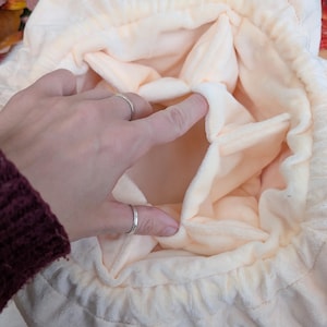 May include: Close-up of a soft, peach-colored drawstring bag with a unique petal-like design. The interior is a lighter shade of peach. The bag is being held open by a hand with silver rings.