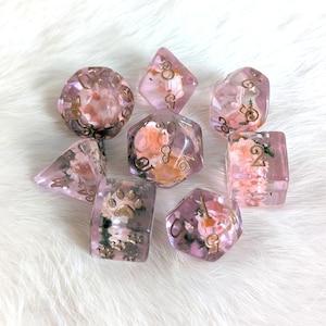 May include: Set of eight pink and gold dice with floral and animal designs. The dice are translucent and have gold numbers and designs.