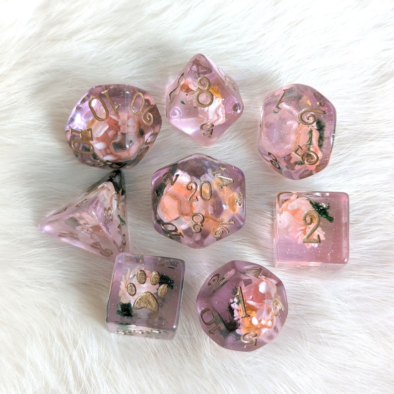 May include: Set of seven pink and clear dice with gold numbers and floral accents. The dice are translucent and show the floral accents inside. The dice are numbered 1-6, 8, 16, and 20.