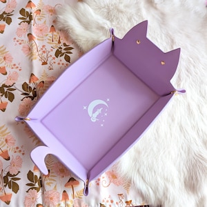 May include: A lavender-colored felt dice tray with a unicorn and crescent moon design. The tray has gold metal accents and is shaped like a cat.