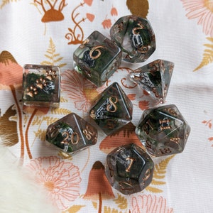 May include: A set of clear resin dice with copper-colored numbers and edges. Each die is filled with green moss and a small, patterned object. The set includes a square die with paw prints and a variety of polyhedral dice.