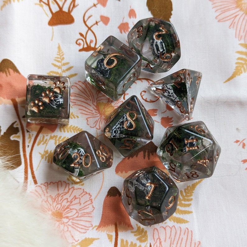 May include: A set of clear resin dice with copper-colored numbers and edges. Each die is filled with green moss and a small, patterned object. The set includes a square die with paw prints and a variety of polyhedral dice.