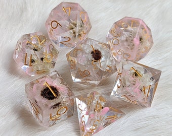 May include: Set of six clear resin dice with gold numbers and dried flowers embedded inside. The dice are in various shapes and sizes, including a d4, d6, d8, d10, d12, and d20.