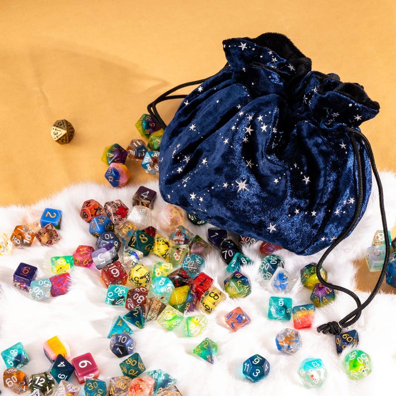 May include: A dark blue velvet drawstring pouch featuring a silver star pattern, accompanied by a large collection of multi-colored polyhedral dice in various shapes and sizes scattered across a white faux fur surface.
