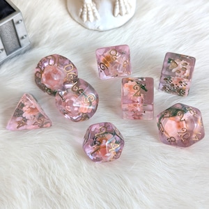 May include: Set of eight pink and gold dice with floral accents. The dice are made of clear resin with pink and gold glitter embedded in the resin. The dice are numbered 1 through 6.
