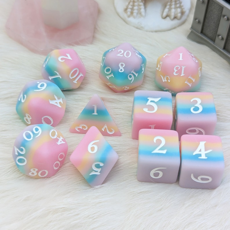May include: A set of 10 pastel-colored dice with rainbow-colored swirls. The dice are made of a soft, opaque material and have white numbers. The dice are arranged in a circle on a white surface.
