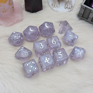 May include: A set of ten purple dice with silver numbers and a moon and star design. The dice are on a white fuzzy surface.