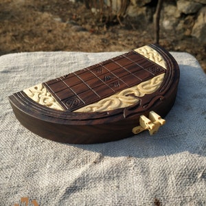 Puede incluir: Un juego de mesa de madera hecho a mano, con una superficie de juego marr&oacute;n oscuro con cuadrados tallados y detalles decorativos de color claro. El tablero del juego tiene forma de barco, con intrincadas tallas en los bordes. Un peque&ntilde;o cierre de color claro asegura el juego.