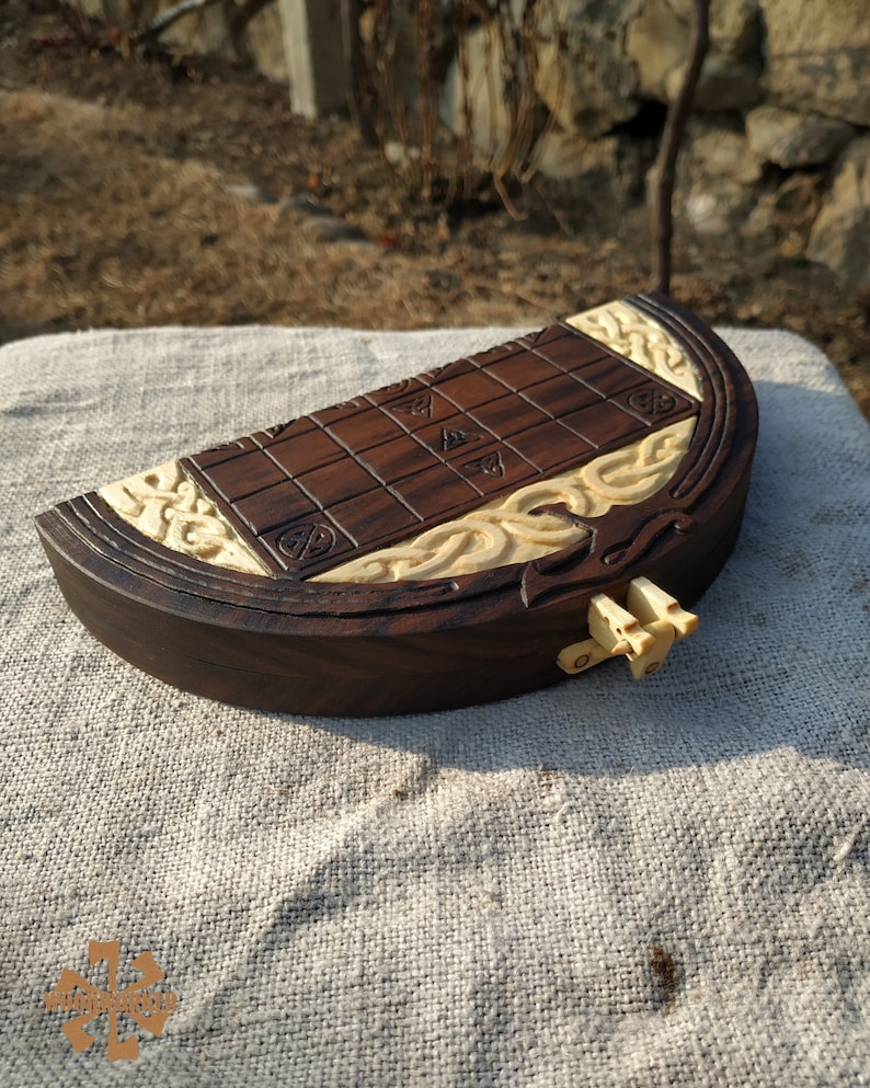 Puede incluir: Un juego de mesa de madera hecho a mano, con una superficie de juego marr&oacute;n oscuro con cuadrados tallados y detalles decorativos de color claro. El tablero del juego tiene forma de barco, con intrincadas tallas en los bordes. Un peque&ntilde;o cierre de color claro asegura el juego.