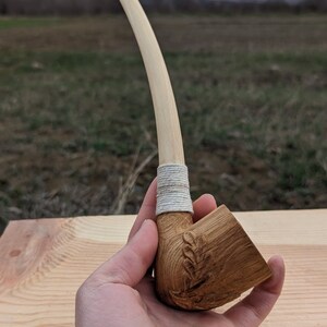 Pu&ograve; includere: Una pipa da fumo in legno con un lungo stelo curvo e una ciotola scolpita con un motivo a foglia. La pipa &egrave; avvolta con uno spago bianco vicino alla ciotola.