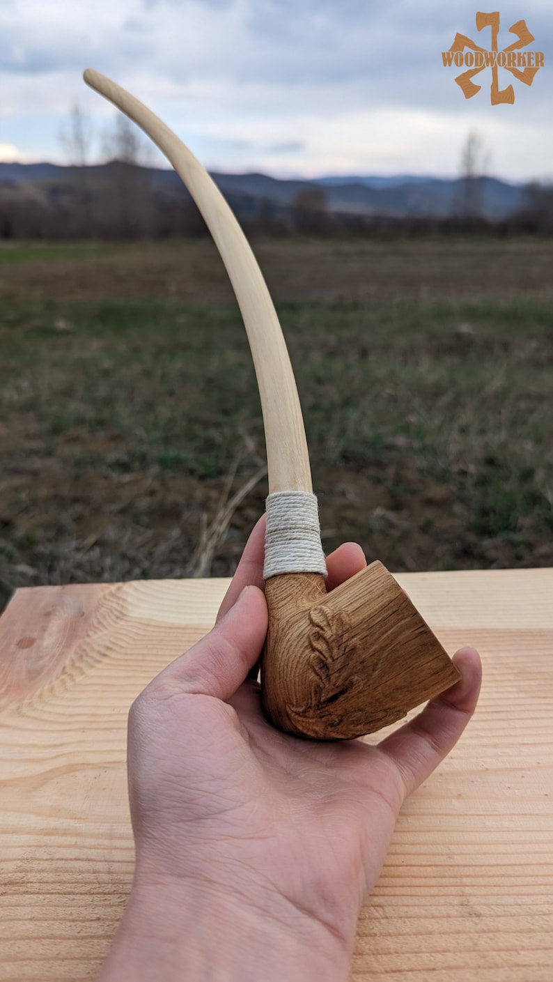 Pu&ograve; includere: Una pipa da fumo in legno con un lungo stelo curvo e una ciotola scolpita con un motivo a foglia. La pipa &egrave; avvolta con uno spago bianco vicino alla ciotola.