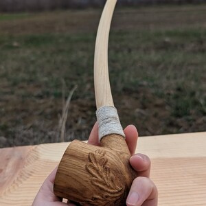 Pu&ograve; includere: Una pipa da fumo in legno con un lungo stelo curvo e una ciotola scolpita con un motivo a foglia di quercia. Lo stelo &egrave; avvolto da un sottile cordoncino bianco.