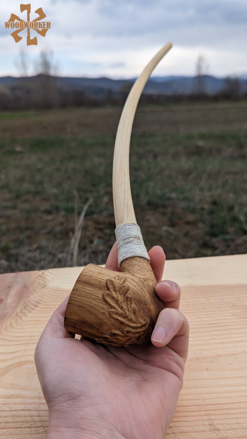 Pu&ograve; includere: Una pipa da fumo in legno con un lungo stelo curvo e una ciotola scolpita con un motivo a foglia di quercia. Lo stelo &egrave; avvolto da un sottile cordoncino bianco.