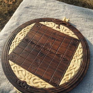 Puede incluir: Un juego de mesa de madera hecho a mano con una superficie de juego marr&oacute;n oscuro y un borde decorativo color crema. El tablero presenta dise&ntilde;os de nudos celtas tallados y s&iacute;mbolos geom&eacute;tricos. El tablero del juego es redondo.