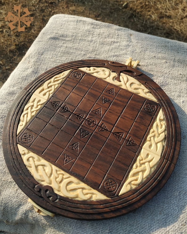 Puede incluir: Un juego de mesa de madera hecho a mano con una superficie de juego marr&oacute;n oscuro y un borde decorativo color crema. El tablero presenta dise&ntilde;os de nudos celtas tallados y s&iacute;mbolos geom&eacute;tricos. El tablero del juego es redondo.