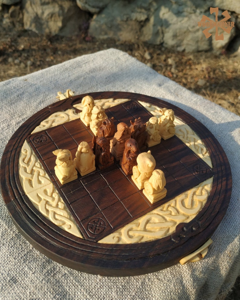 Puede incluir: Un juego de ajedrez de madera hecho a mano con piezas talladas intrincadamente y un tablero redondo. El tablero presenta una superficie de juego marr&oacute;n oscuro con detalles tallados de color claro y un borde decorativo. Las piezas de ajedrez son en tonos de madera claros y oscuros.