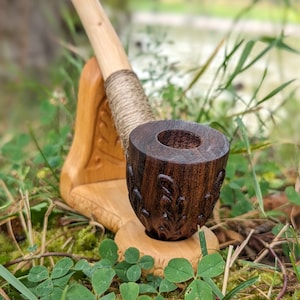 May include: A wooden pipe with a carved bowl and a long, curved stem. The pipe is resting on a wooden stand with a carved design. The pipe and stand are surrounded by green grass and moss.