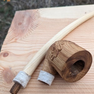 Pu&ograve; includere: Una pipa da fumo in legno con un design intagliato sulla ciotola. La pipa ha un gambo lungo e sottile con una corda bianca avvolta attorno all'estremit&agrave;. La pipa &egrave; appoggiata su una superficie di legno.