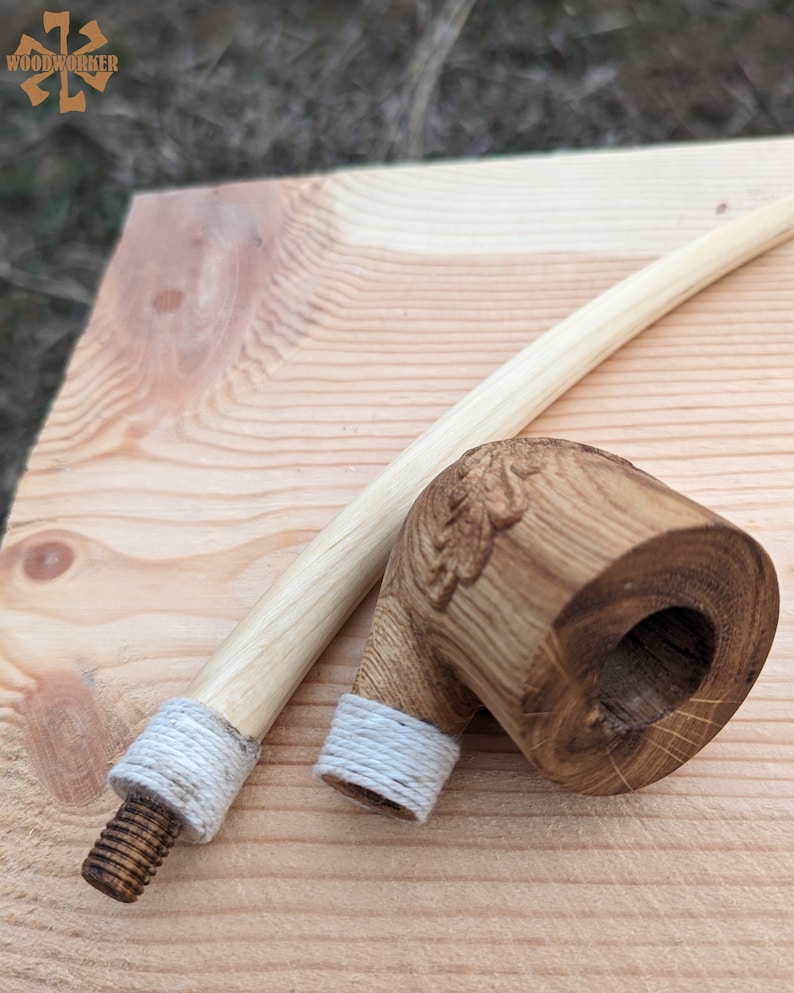 Pu&ograve; includere: Una pipa da fumo in legno con un design intagliato sulla ciotola. La pipa ha un gambo lungo e sottile con una corda bianca avvolta attorno all'estremit&agrave;. La pipa &egrave; appoggiata su una superficie di legno.