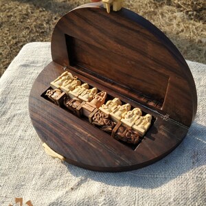 Puede incluir: Un juego de ajedrez de madera con una caja circular marr&oacute;n oscuro. Las piezas del ajedrez est&aacute;n talladas en color beige claro y se guardan en un compartimento ajustado. La tapa est&aacute; abierta, revelando las piezas del ajedrez. El juego est&aacute; sobre una superficie de color claro.