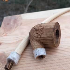 Pu&ograve; includere: Una pipa da fumo in legno con un motivo a foglia intagliato e una corda bianca avvolta attorno al gambo. La pipa &egrave; realizzata in legno marrone chiaro e ha una finitura naturale.