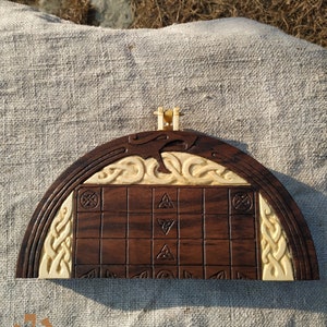 Puede incluir: Un juego de mesa de madera hecho a mano con una superficie de juego marr&oacute;n oscuro y detalles tallados de color crema. El tablero presenta un patr&oacute;n de cuadr&iacute;cula con s&iacute;mbolos tallados y bordes decorativos, y un peque&ntilde;o asa en la parte superior.