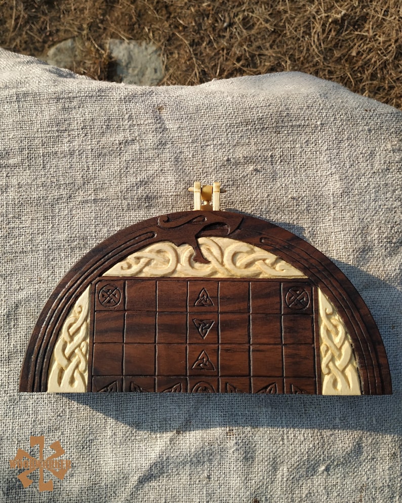 Puede incluir: Un juego de mesa de madera hecho a mano con una superficie de juego marr&oacute;n oscuro y detalles tallados de color crema. El tablero presenta un patr&oacute;n de cuadr&iacute;cula con s&iacute;mbolos tallados y bordes decorativos, y un peque&ntilde;o asa en la parte superior.