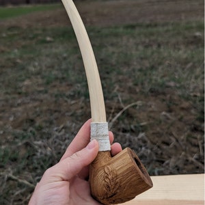 Pu&ograve; includere: Una pipa da fumo in legno con un lungo stelo curvo. La ciotola della pipa &egrave; intagliata con un motivo a foglia ed &egrave; tenuta in una mano. La pipa &egrave; realizzata in legno marrone chiaro e ha una corda bianca avvolta attorno allo stelo.