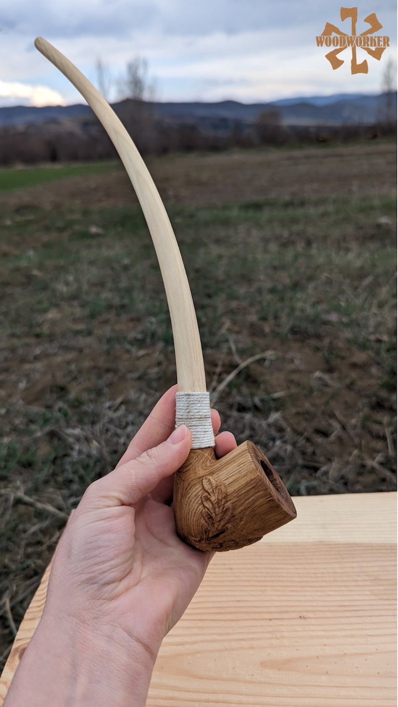 Pu&ograve; includere: Una pipa da fumo in legno con un lungo stelo curvo. La ciotola della pipa &egrave; intagliata con un motivo a foglia ed &egrave; tenuta in una mano. La pipa &egrave; realizzata in legno marrone chiaro e ha una corda bianca avvolta attorno allo stelo.