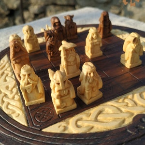 Puede incluir: Un juego de ajedrez de madera con piezas talladas. El tablero es marr&oacute;n oscuro con tallas crema. Las piezas representan figuras vikingas. El logotipo "WOODWORKER" es visible en la esquina superior derecha.