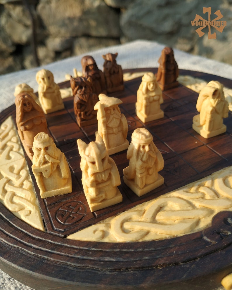 Puede incluir: Un juego de ajedrez de madera con piezas talladas. El tablero es marr&oacute;n oscuro con tallas crema. Las piezas representan figuras vikingas. El logotipo "WOODWORKER" es visible en la esquina superior derecha.
