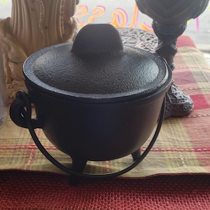 May include: A black cast iron cauldron with a lid, three small legs, and a handle. The cauldron sits on a red and green plaid tablecloth. The cauldron is a dark, matte black color.