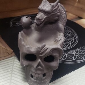 Puede incluir: Un quemador de incienso de cerámica negra con forma de cráneo con un dragón en la parte superior. El dragón sostiene una pequeña bola en su boca.