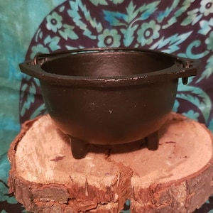May include: A small, black cast iron cauldron with three short legs and a handle. The cauldron is sitting on a round, natural wood slice. The background features a teal and navy floral pattern.