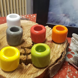 Könnte beinhalten: Eine Sammlung farbenfroher Zahnbürstenhalter aus Keramik in verschiedenen Farben, darunter Schwarz, Weiß, Grau, Rot, Orange, Gelb und Grün. Sie sind auf einer Holzscheibe angeordnet, mit Kristallen im Hintergrund.