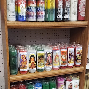 Puede incluir: Una estantería de madera exhibe una variedad de velas en frascos de vidrio. Las velas presentan etiquetas coloridas con figuras religiosas y texto, incluyendo "ORULA", "ELEGU" y "OYA". Las velas son de varios colores, incluyendo blanco, rojo, azul y verde.