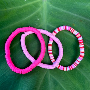May include: Three pink and white beaded bracelets with different patterns. The bracelets are on a green leaf background.