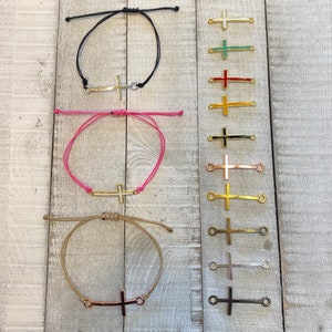 May include: Three adjustable bracelets with different colored cords and a silver cross charm. The cords are black, pink, and beige. The cross charms are all the same size and shape. There are also 10 different colored cross charms displayed below the bracelets.