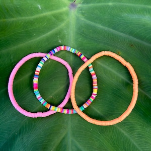 May include: Three colorful bracelets made of small, round beads. The bracelets are pink, orange, and multi-colored. The bracelets are arranged in a circle on a green leaf.
