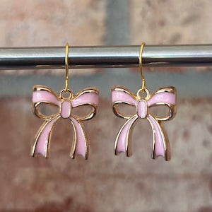 May include: A pair of gold earrings with pink bow charms. The bows are detailed with gold trim.