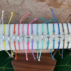 May include: A collection of 20 colorful string bracelets with a silver cross charm. The bracelets are displayed on a wooden surface.