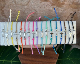 May include: A collection of 20 colorful string bracelets with a silver cross charm. The bracelets are displayed on a wooden surface.